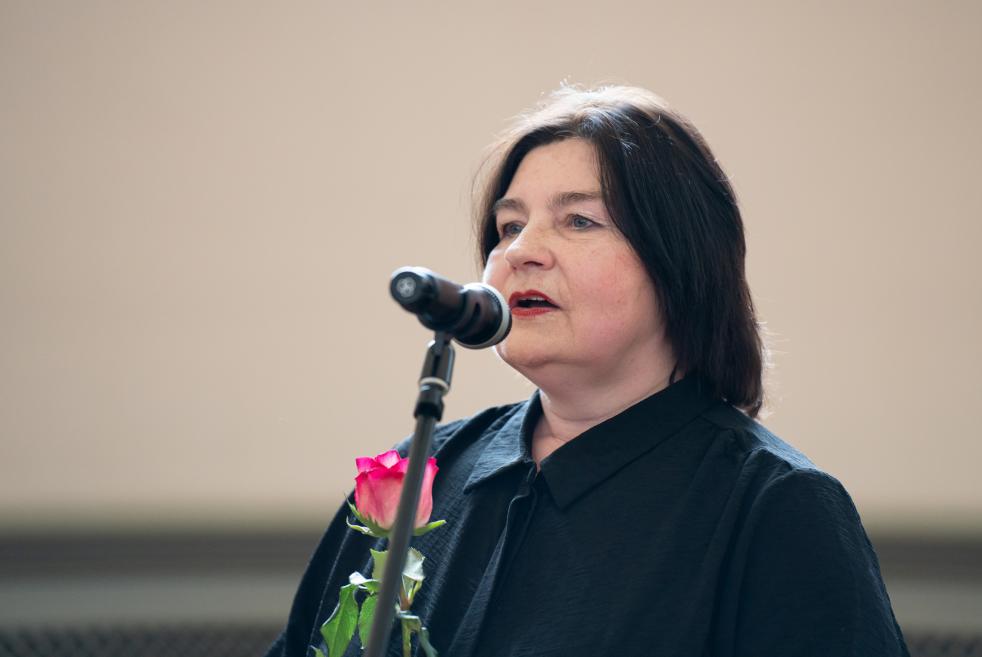 Sieviete pie mikrofona runā, rokā turot rozes ziedu.