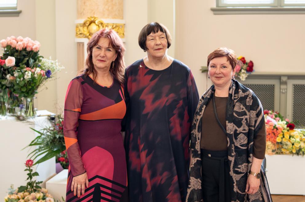 Trīs sievietes stāv kopā un pozē foto muzeja zālē, apkārt redzami ziedu pušķi.