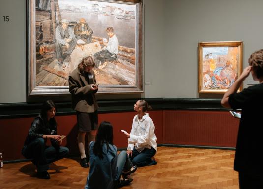 Muzeja zālē pie sienas ar gleznām vairāki jaunieši sēž un stāv uz grīdas, sarunājas un skatās piezīmes, kamēr viens apmeklētājs aplūko darbus.