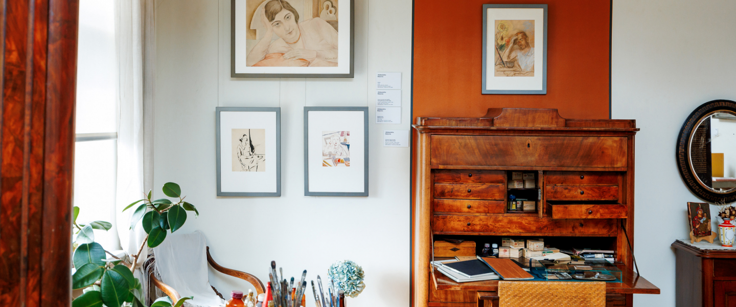 In a bright interior, a workspace features a wooden desk with art materials, drawings displayed on the wall, and a houseplant nearby.