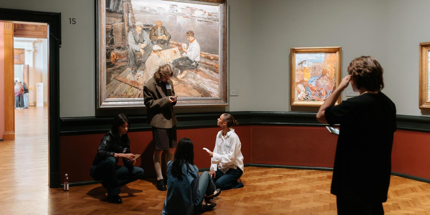 Muzeja zālē pie sienas ar gleznām vairāki jaunieši sēž un stāv uz grīdas, sarunājas un skatās piezīmes, kamēr viens apmeklētājs aplūko darbus.