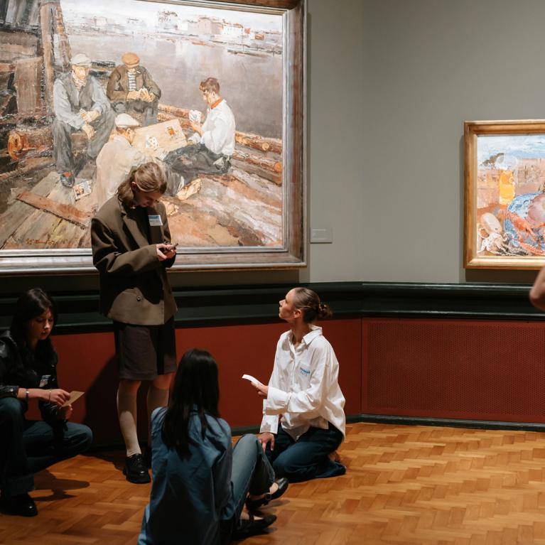 Muzeja zālē pie sienas ar gleznām vairāki jaunieši sēž un stāv uz grīdas, sarunājas un skatās piezīmes, kamēr viens apmeklētājs aplūko darbus.