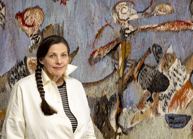 Sieviete ar bizi stāv muzeja telpā abstrakta gobelēna priekšā, kas veidots zilganos, brūnos un sarkanīgos toņos ar figurāliem motīviem.