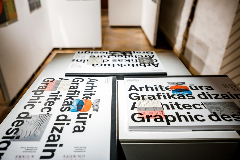 Uz galda izlikti izstādes katalogi ar uzrakstu “Arhitektūra. Grafikas dizains. Architecture. Graphic design.”.