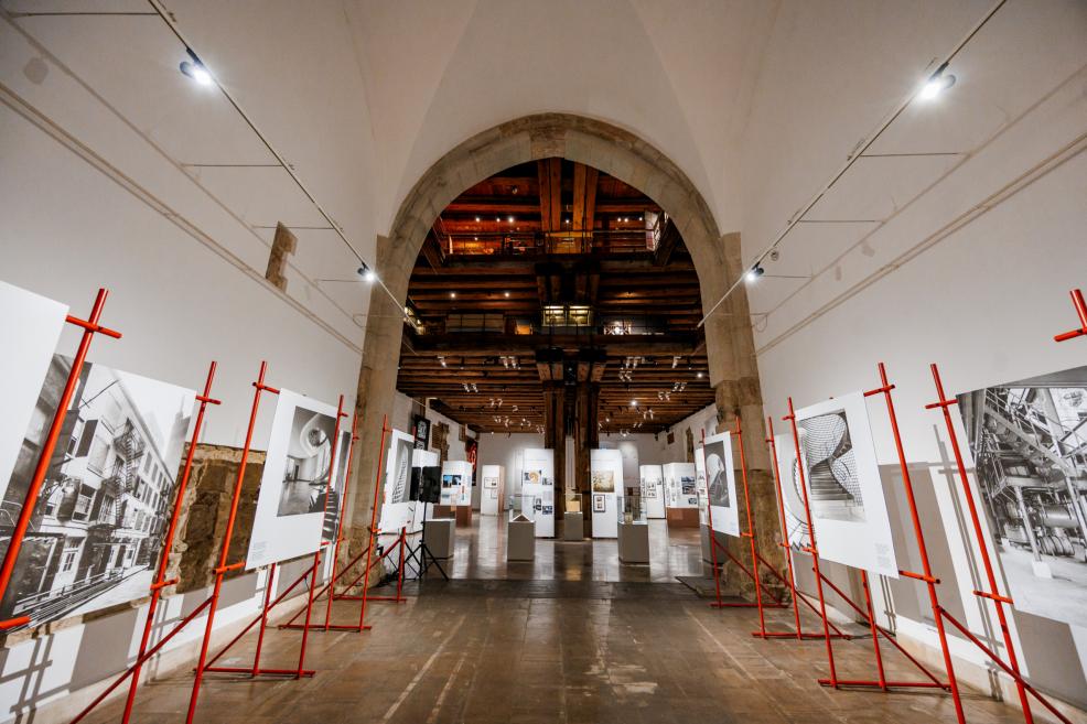 Izstādes zāle ar arhitektūras un dizaina ekspozīciju, skatāmas lielas melnbaltas fotogrāfijas uz sarkaniem statīviem.