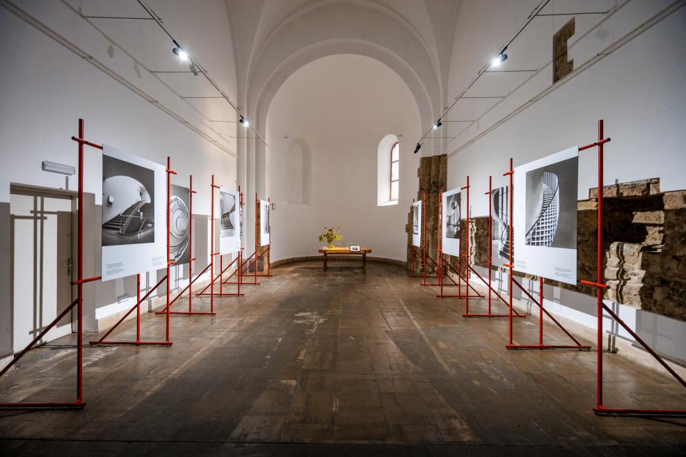 Izstādes gaitenis ar sarkaniem metāla rāmjiem, kuros izvietotas lielizmēra melnbaltas arhitektūras fotogrāfijas.