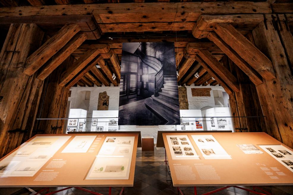 Izstādes skats ar arhitektūras zīmējumiem un lielu karājošu kāpņu fotogrāfiju zem koka sijām.