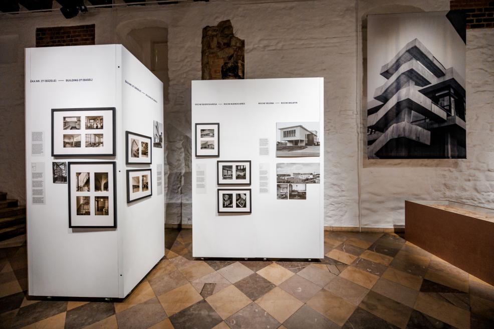Izstādes paneļi ar vēsturiskām melnbaltām fotogrāfijām un arhitektūras plāniem uz baltiem stendiem.
