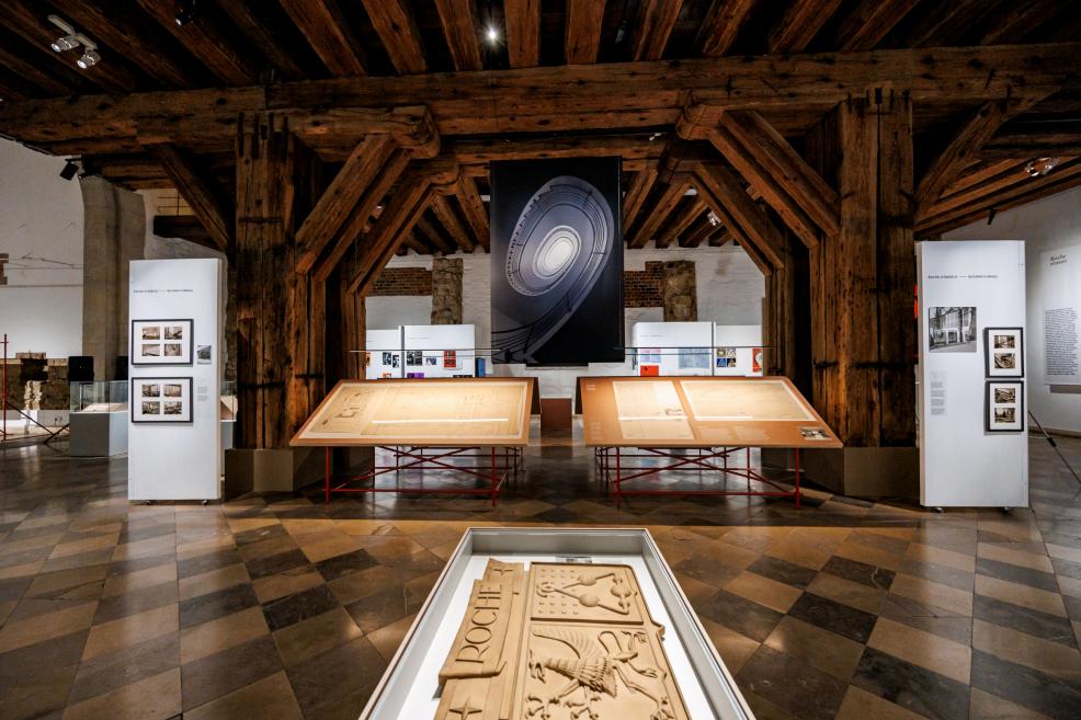 Tuvplāns ar lieliem arhitektūras rasējumiem, kas eksponēti uz horizontāliem stendiem Roche izstādē.