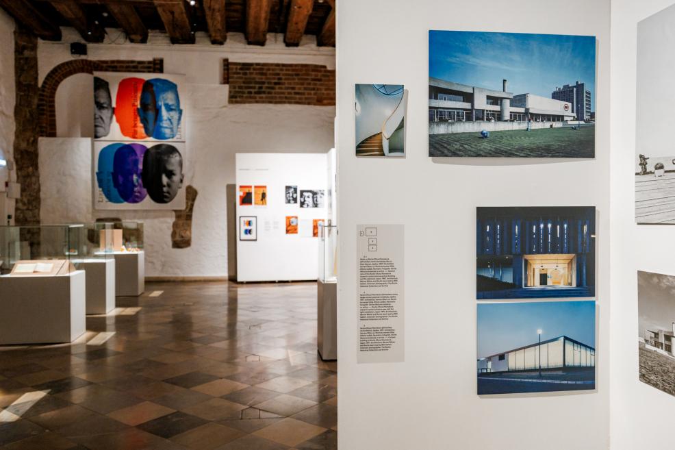 Izstādes paneļi ar modernisma ēku fotogrāfijām un fona sienā redzami krāsaini plakāti.