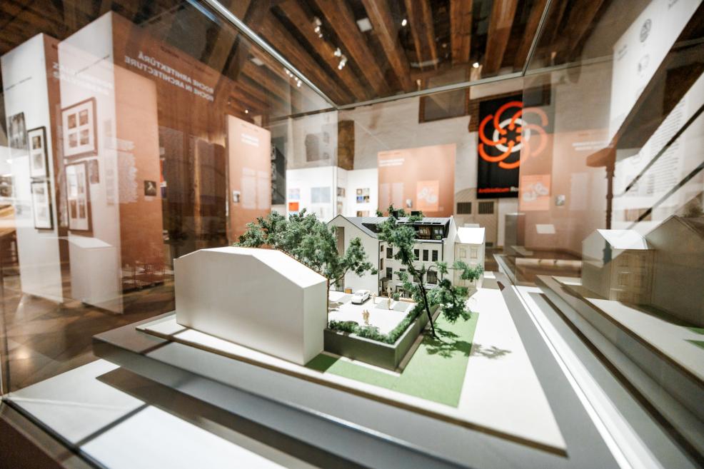 Arhitektūras makets stikla vitrīnā ar kokiem un ēkām.