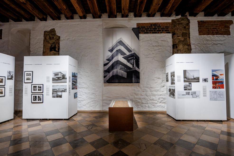 Izstādes paneļi ar Roche ēku fotogrāfijām un plāniem, eksponēti uz baltiem stendiem pie mūra sienas.