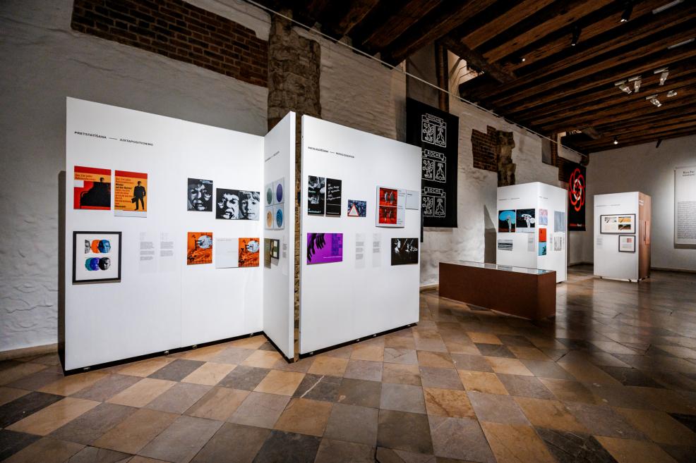 Izstādes ekspozīcija ar grafikas darbiem un krāsainiem plakātiem uz baltiem stendiem.