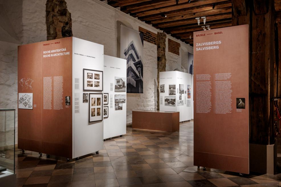 Izstādes skats ar tekstu paneļiem un fotogrāfijām par Roche arhitektūru zālē ar mūra sienām un koka griestiem.
