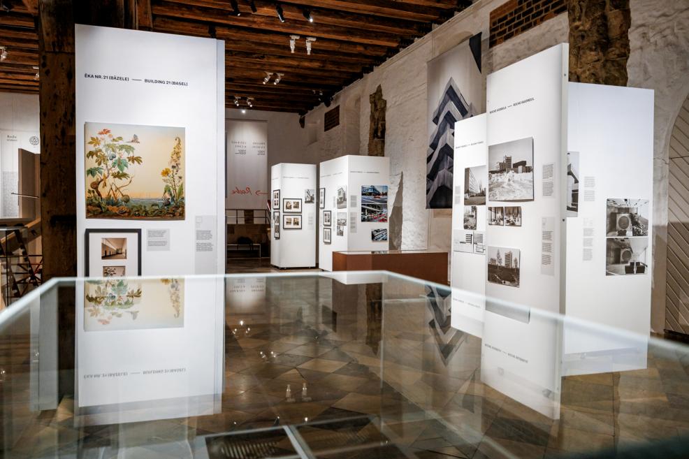 Izstādes skats ar baltiem paneļiem, kuros redzami Roche arhitektūras attēli, fonā vēsturiska ēkas fotogrāfija.