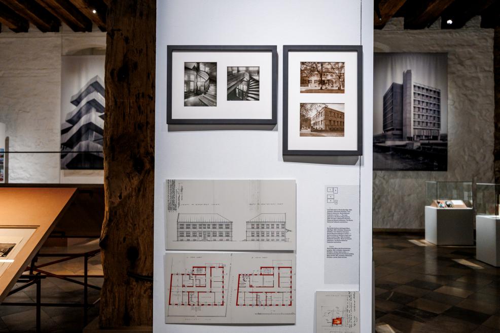 Izstādes ekspozīcija ar vēsturiskām fotogrāfijām un ēkas plāniem uz sienas.