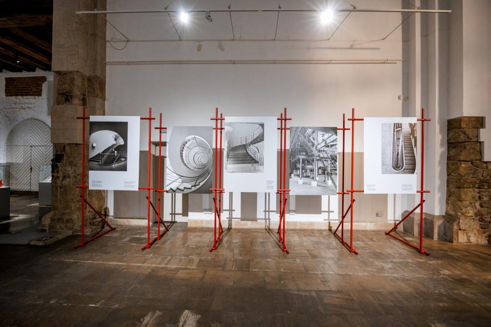 Izstādes skats ar melnbaltām arhitektūras fotogrāfijām, kas piestiprinātas pie sarkaniem stendiem telpā ar mūra sienām.
