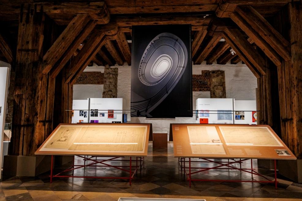 Tuvplāns ar lieliem arhitektūras rasējumiem, kas eksponēti uz horizontāliem stendiem Roche izstādē.