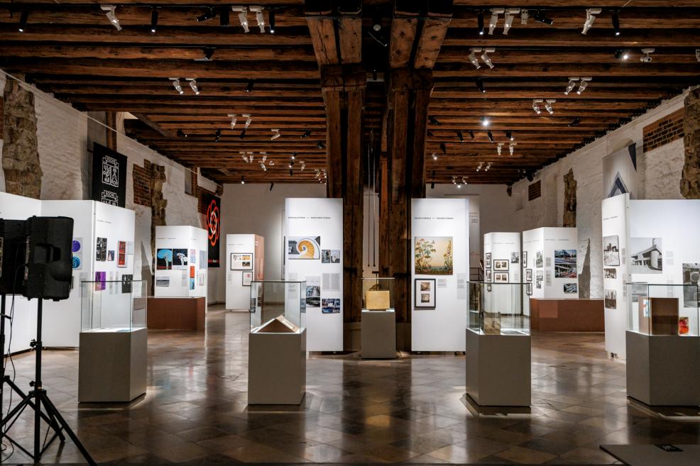 Plaša izstādes telpa ar vitrīnām un stendiem, kuros redzami arhitektūras un mākslas darbi.