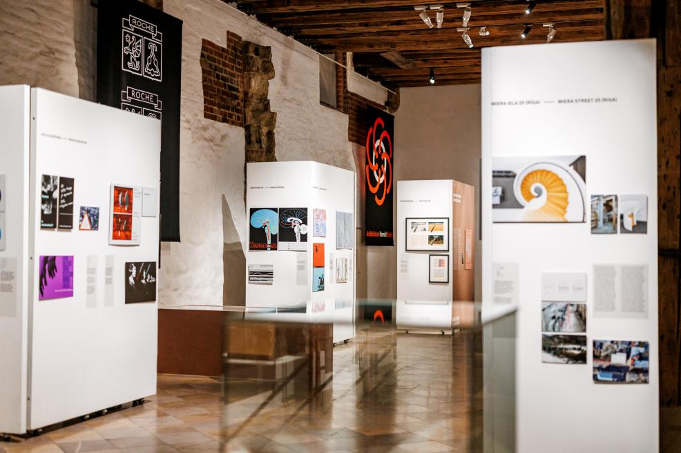 Izstādes ekspozīcijas fragmentā redzamas grafiskā dizaina plakātu kompozīcijas uz baltiem paneļiem.