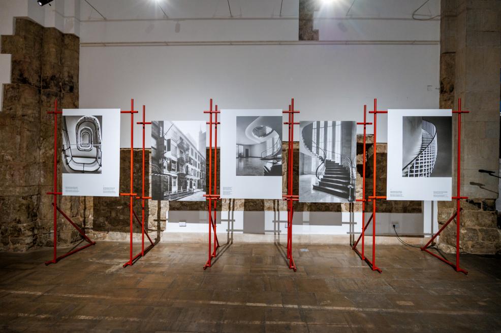 Izstādes instalācija ar lielizmēra melnbaltām arhitektūras fotogrāfijām, eksponētām uz sarkaniem metāla stendiem.