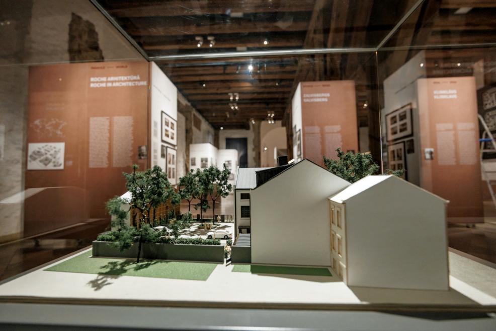 Izstādes skats ar arhitektūras maketu vitrīnā, attēlojot ēku ar dārzu, fonā tekstu paneļi.