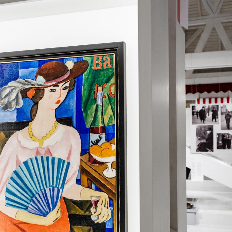 Muzeja ekspozīcijā redzama krāsaina glezna ar sievieti cepurē, kura tur vēdekli un sēž pie galda ar augļiem un dzērienu, fonā – citas izstādes fotogrāfijas.