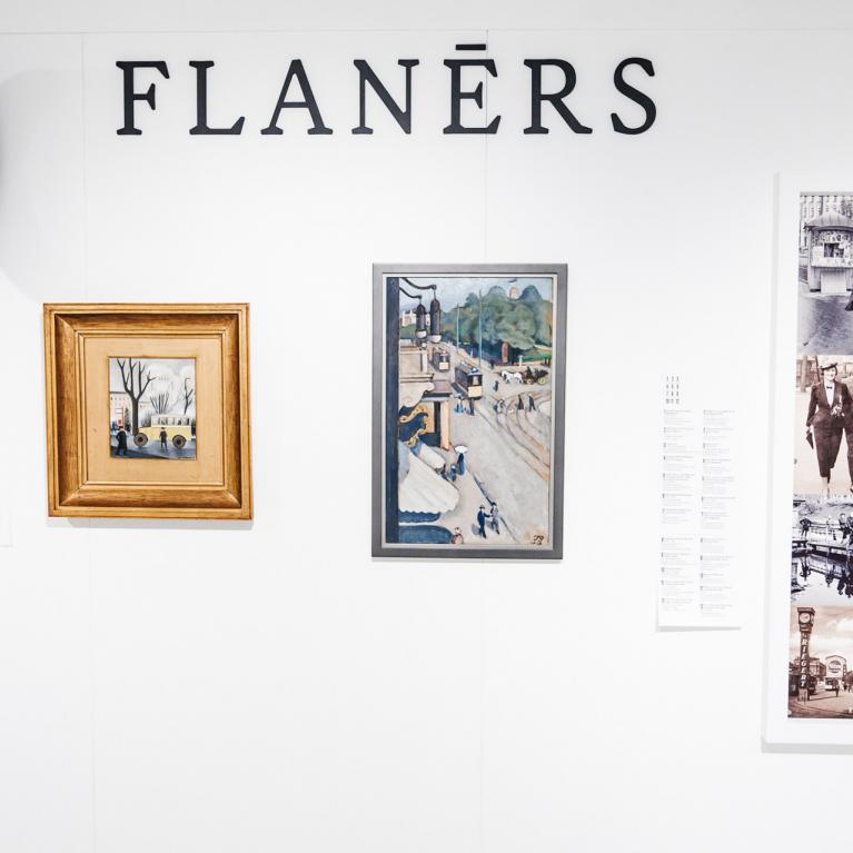 Muzeja sienas ekspozīcija ar uzrakstu “FLANĒRS”, redzamas gleznas, vēsturiskas fotogrāfijas un sienas pulkstenis.