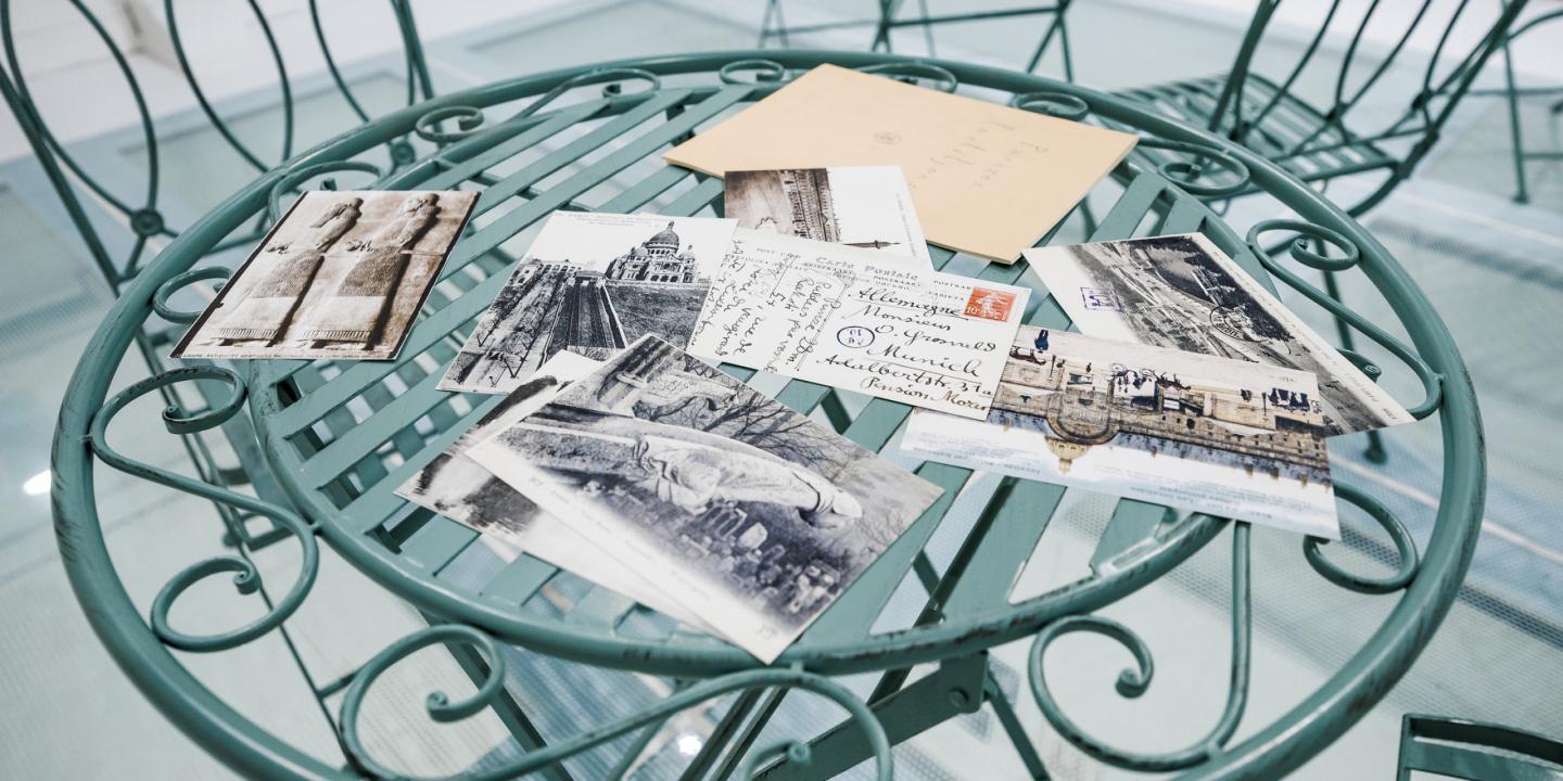 Uz apaļa metāla galdiņa izklātas vēsturiskas pastkartes un aploksne ar rokrakstu, redzami melnbalti pilsētas skati.