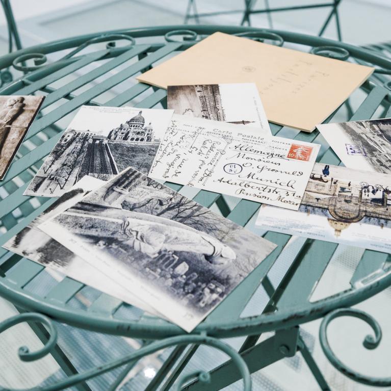 Uz apaļa metāla galdiņa izklātas vēsturiskas pastkartes un aploksne ar rokrakstu, redzami melnbalti pilsētas skati.