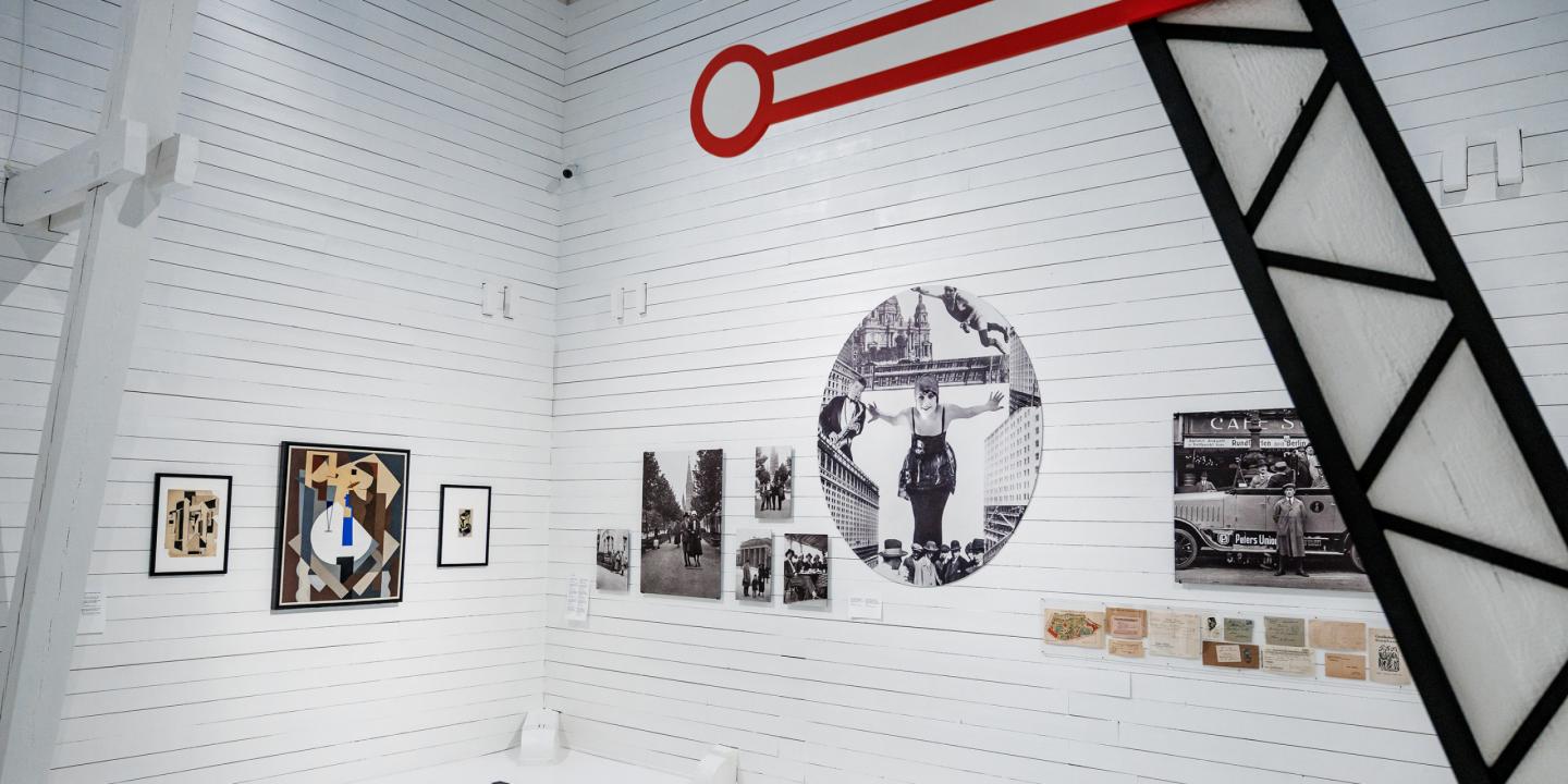 Muzeja ekspozīcija baltā zālē ar modernisma gleznām, vēsturiskām fotogrāfijām un dokumentiem, virs sienas redzams grafisks sarkanas atslēgas motīvs.