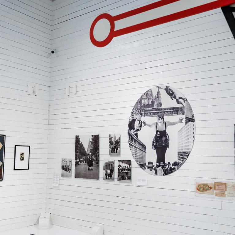 Muzeja ekspozīcija baltā zālē ar modernisma gleznām, vēsturiskām fotogrāfijām un dokumentiem, virs sienas redzams grafisks sarkanas atslēgas motīvs.