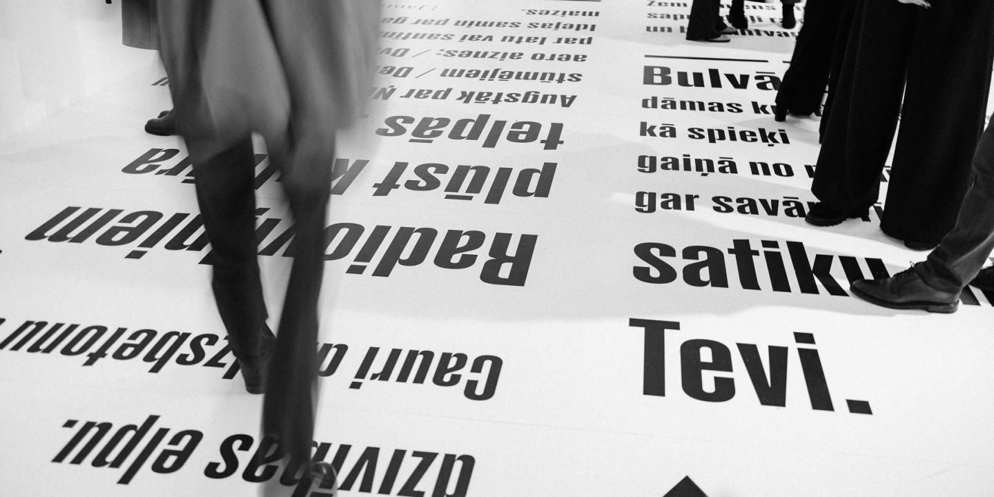 Melnbalts skats ar cilvēkiem, kas staigā pa grīdu, uz kuras lieliem burtiem drukāti latviešu valodas teksti.