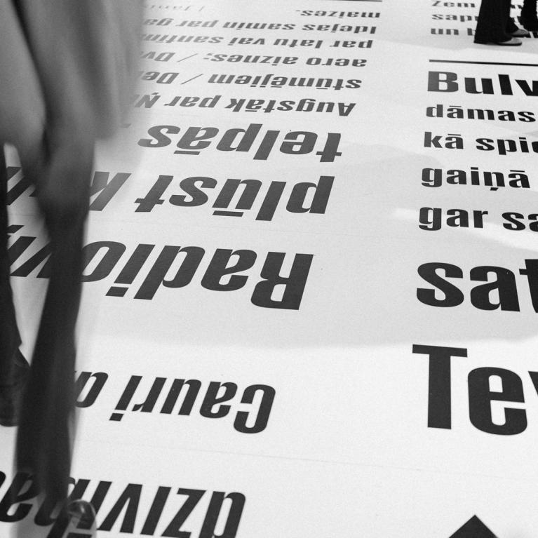 Melnbalts skats ar cilvēkiem, kas staigā pa grīdu, uz kuras lieliem burtiem drukāti latviešu valodas teksti.