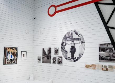 Muzeja ekspozīcija baltā zālē ar modernisma gleznām, vēsturiskām fotogrāfijām un dokumentiem, virs sienas redzams grafisks sarkanas atslēgas motīvs.