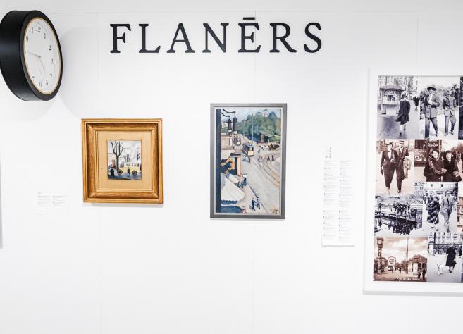 Muzeja sienas ekspozīcija ar uzrakstu “FLANĒRS”, redzamas gleznas, vēsturiskas fotogrāfijas un sienas pulkstenis.