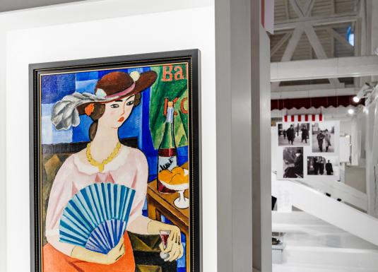 Muzeja ekspozīcijā redzama krāsaina glezna ar sievieti cepurē, kura tur vēdekli un sēž pie galda ar augļiem un dzērienu, fonā – citas izstādes fotogrāfijas.