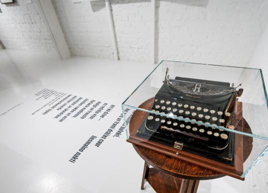 Muzeja ekspozīcijā uz neliela galdiņa zem stikla pārsega novietota vēsturiska rakstāmmašīna, bet uz grīdas redzams tipogrāfisks teksta fragments.