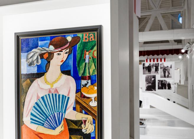 Muzeja ekspozīcijā redzama krāsaina glezna ar sievieti cepurē, kura tur vēdekli un sēž pie galda ar augļiem un dzērienu, fonā – citas izstādes fotogrāfijas.