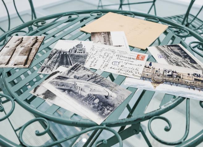 Uz apaļa metāla galdiņa izklātas vēsturiskas pastkartes un aploksne ar rokrakstu, redzami melnbalti pilsētas skati.