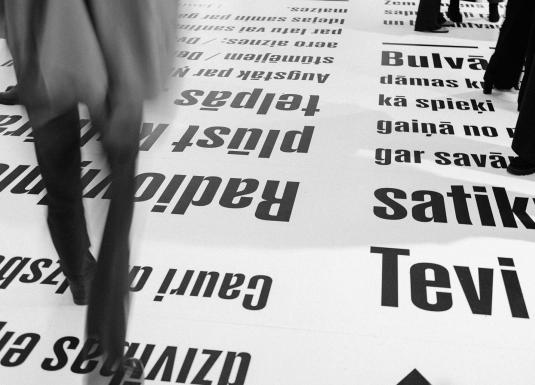 Melnbalts skats ar cilvēkiem, kas staigā pa grīdu, uz kuras lieliem burtiem drukāti latviešu valodas teksti.