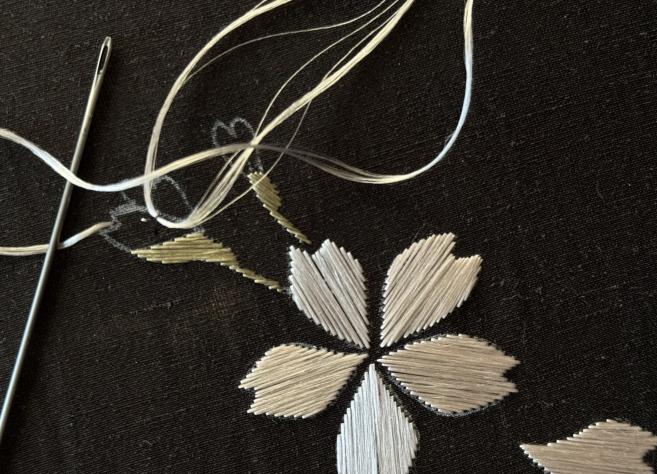 Tekstila izšuvuma detaļa ar adatu un diegu uz tumša auduma.