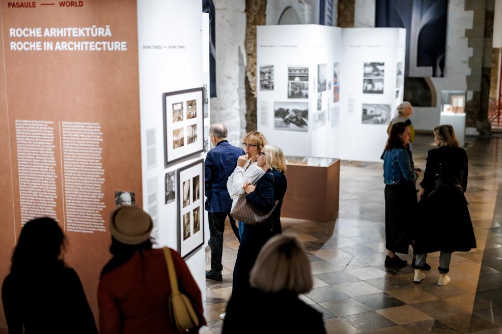 People at the exhibition opening event are conversing and viewing the displays, with flowers visible in the foreground.