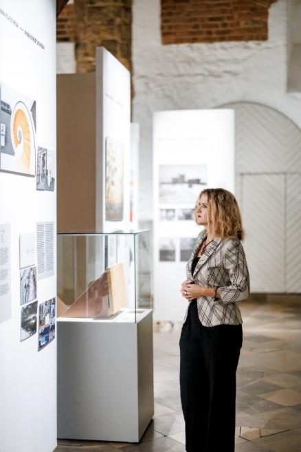 Sieviete muzeja izstādē skatās uz ekspozīcijas sienu ar fotogrāfijām un tekstiem.