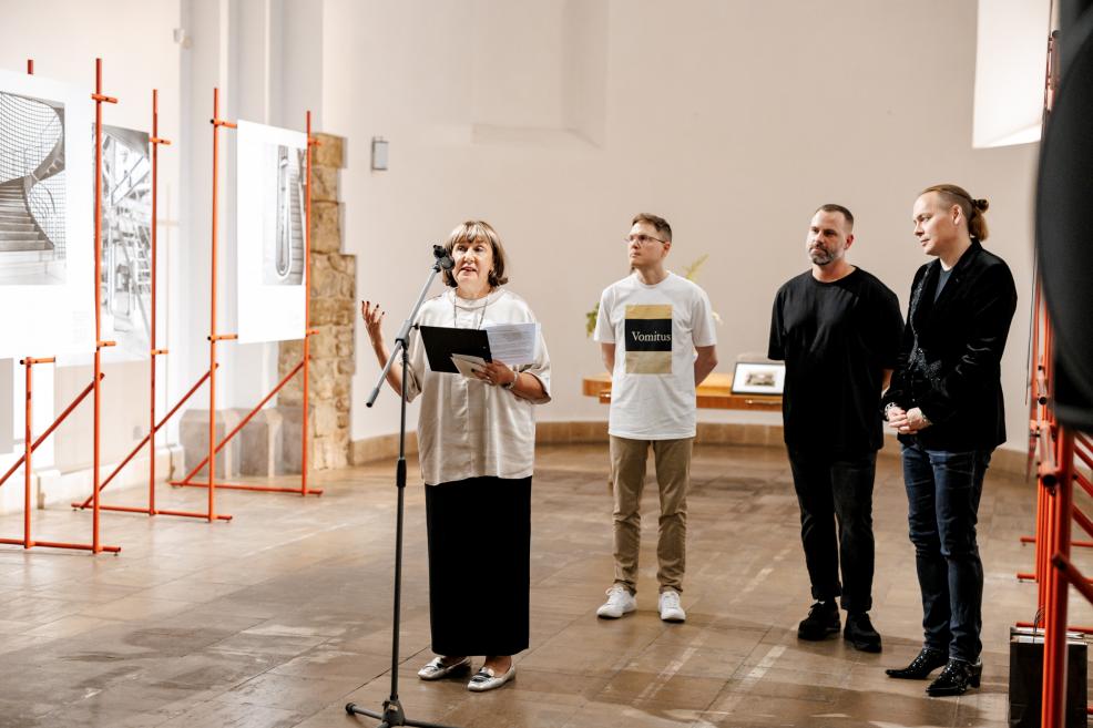 Sieviete runā pie mikrofona izstādes atklāšanā, aiz viņas stāv kolēģi pie arhitektūras fotogrāfijām.