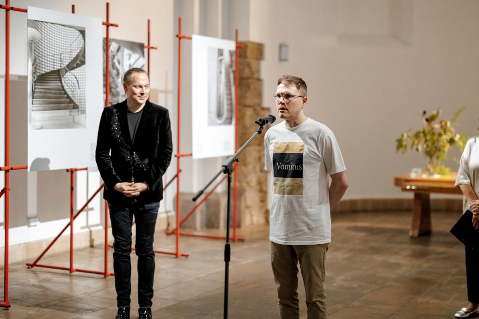 Vīrietis pie mikrofona uzstājas izstādes atklāšanā, fonā arhitektūras fotogrāfijas.