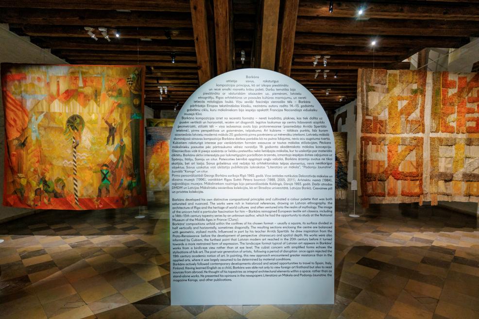 Izstādes ekspozīcijas skats ar tekstilmākslas darbiem un informatīvu apraksta stendu priekšplānā.