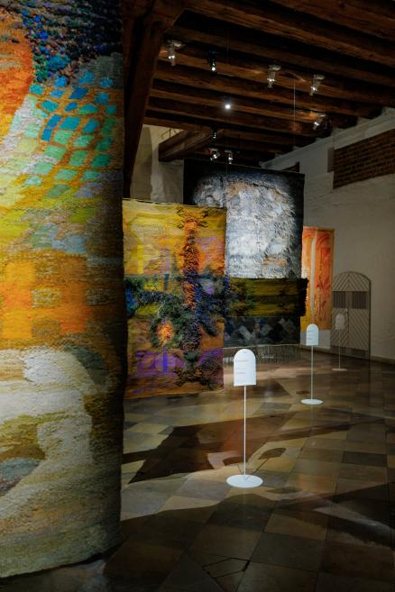 Tekstilmākslas darbu ekspozīcijas skats ar vairākiem liela formāta, reljefiem tekstiliju darbiem dažādos zemes, dzeltenos un tumšos toņos muzeja vēsturiskajā interjerā.