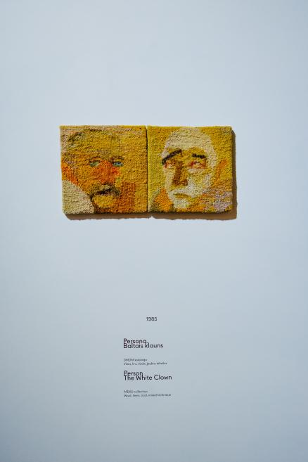 Tekstilmākslas darbs – divdaļīgs portrets siltos dzeltenos un oranžos toņos, kurā attēlotas divas stilizētas sejas, eksponēts uz gaišas muzeja sienas ar darba anotāciju zem tā.