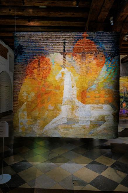 Muzeja zālē piekārts liela formāta gobelēns siltos dzeltenos, oranžos un baltos toņos ar stilizētu cilvēka figūru un arhitektonisku formu siluetiem.